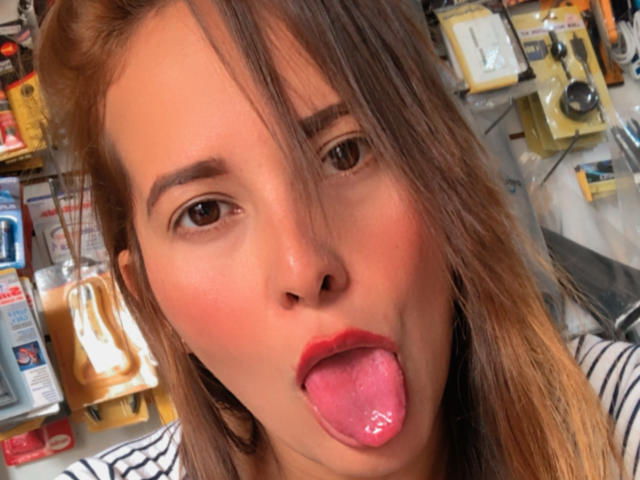 SaritaVelez has light brown skin, red lipstick, and brown eyes, with shoulder-length brown hair in a messy style. She's wearing a striped shirt, looking at the camera.