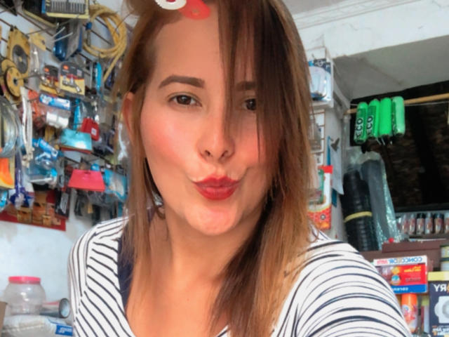SaritaVelez with her tongue out, sticking it up, showing her pink tongue and red lips, brown eyes, and light brown hair with a slight side-swept look.