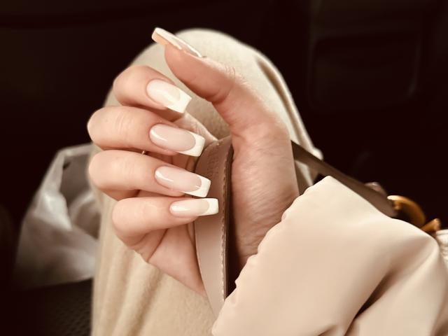 SugarrX's hand with long, almond-shaped, nude-pink acrylic nails, holding a brown card, wearing a beige jacket, fingers slightly curled.