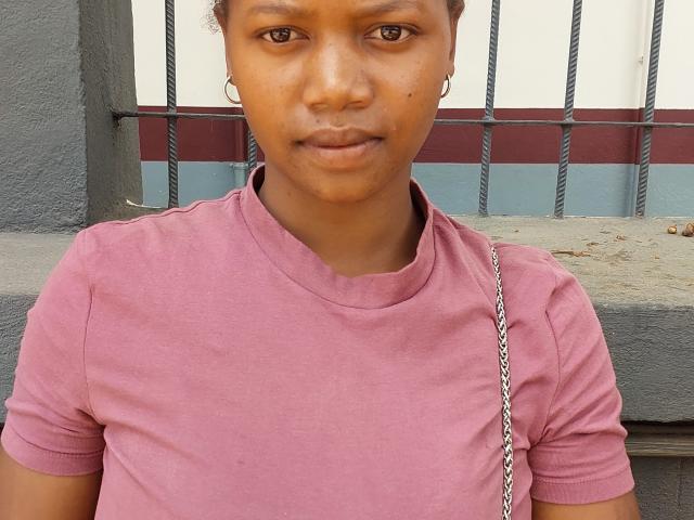 CarinahVanilla, brown-skinned with short curly hair, wearing a pink t-shirt, silver chain, and hoop earrings, has a serious expression and small nose. Her pussy is not visible.