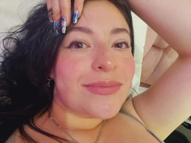 VaaleryStark with pink cheeks, nose piercing, and black hair, smiling with hand on head, wearing colorful nail polish, and a gray top, showing a cute, relaxed face.