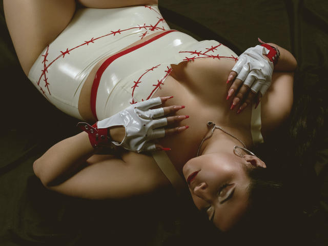 KamileNilsen lying down, wearing a white latex bodysuit with red thorns, white gloves with red accents, and a necklace, with hands on chest, showing off her tits.