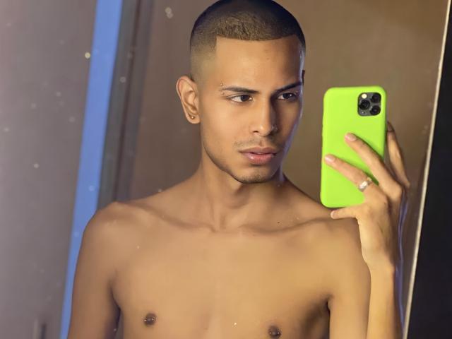 TybelMont, shirtless, holding a neon green phone, taking a mirror selfie. He has a short buzz cut, nose piercing, and small nipples. His expression is neutral, looking at the camera.