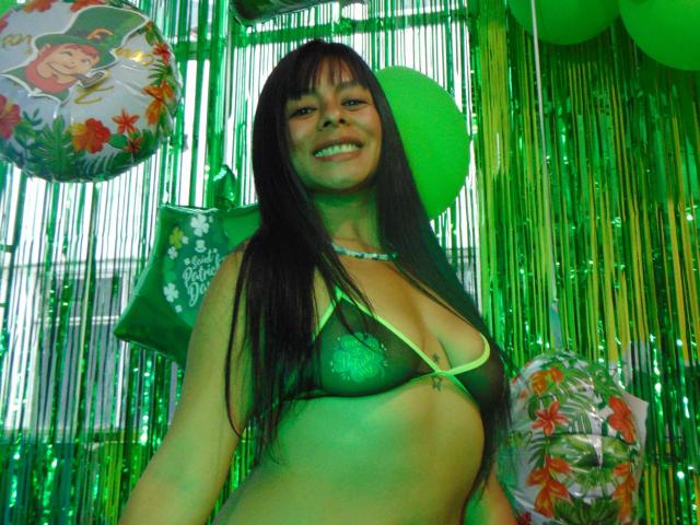 SoffyChasse, with long black hair and green bra, has small tits, a necklace, and a greenish glow, smiling at the camera, surrounded by green balloons and streamers.