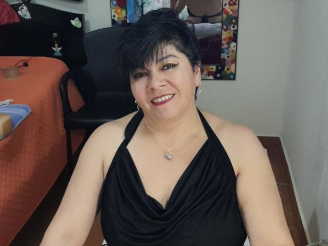 angellovelly, short black hair, smiling, wearing black halter top, showing small tits, necklace with pearl, sitting in chair, wooden floor, orange bed cover in background.