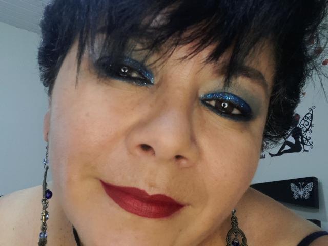 Angellovelly has dark, curly hair, blue glitter eyeshadow, and red lipstick. She's wearing purple earrings and has a slight smile with her nose in the center.