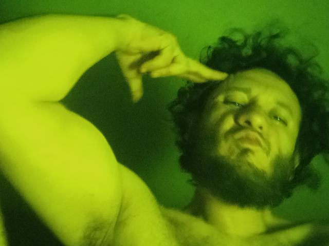 DavydWolfy69, senza camicia, con capelli scuri ricci e barba, indica la fronte con la mano destra, viso e braccio con una sfumatura verde, leggermente sfocato, guarda verso l'alto con un'espressione rilassata..