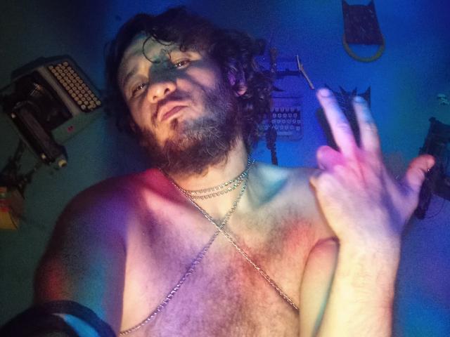 DavydWolfy69, shirtless, curly brown hair, beard, chain necklace, holding cigarette, blue and purple light, chest hair, slightly annoyed expression, right hand raised, left arm partially visible.