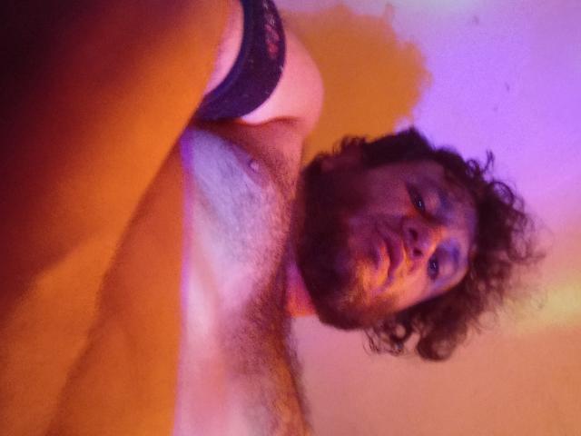 DavydWolfy69, sem camisa, com cabelo castanho encaracolado, olhos azuis e barba, usando uma manga de renda preta, em um selfie borrado, colorido e com pouca luz, mostrando o peito peludo..
