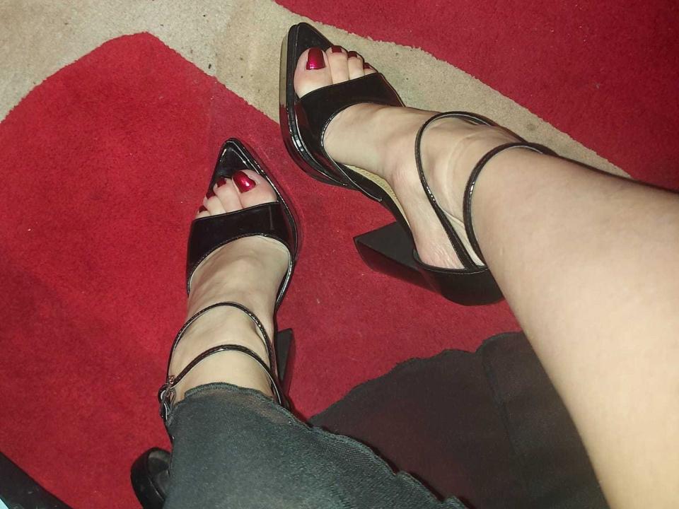 DiabolicaDiva's legs in black high heels with shiny pointed toes, red-painted toenails, and black strappy sandals, wearing dark jeans, on a red carpet.