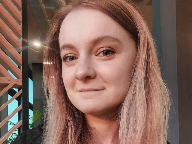 AlmaFlowers heeft lichtroze-bruin haar, een bleke huid en een kleine glimlach, met subtiele eyeliner en een licht glanzende neus. Haar ogen zijn iets samengeknepen, en ze ziet er ontspannen uit..