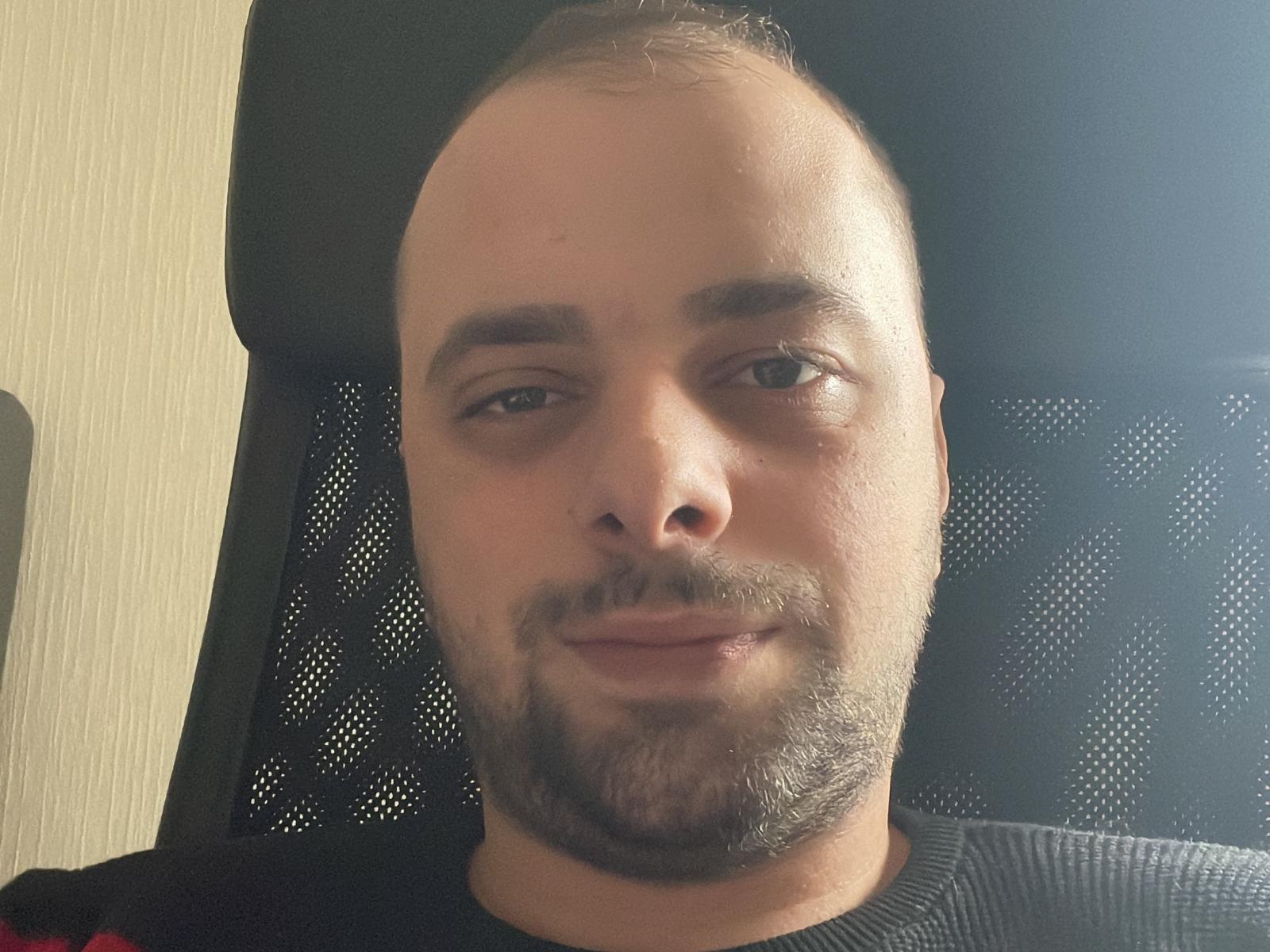JessHood ha la testa calva, una barba marrone chiaro e sta seduto su una sedia nera in rete, indossando un maglione scuro. Sta sorridendo, con un leggero sollevamento del mento..