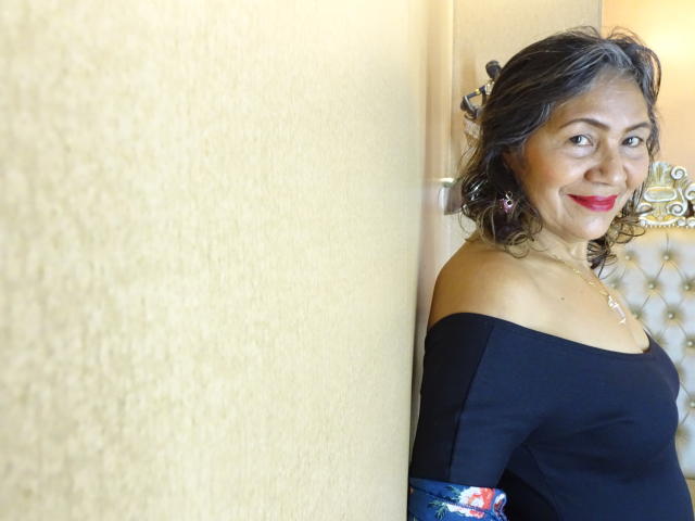CelesteBakers, middle-aged woman with curly brown hair, red lipstick, black off-shoulder top, smiling, standing against a beige wall, gold jewelry, white tufted background.