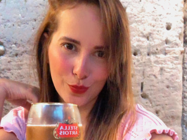 SaritaVelez with light brown hair, red lipstick, holding a beer glass, pink shirt, slightly smiling, and making a kissing gesture with her lips.