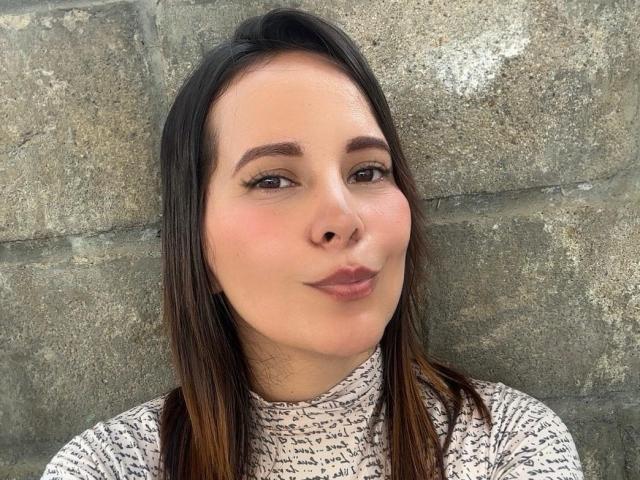 SaritaVelez with brown hair, light skin, and pink lips, wearing a white turtleneck with black script, pouting slightly. She's against a rough stone wall.
