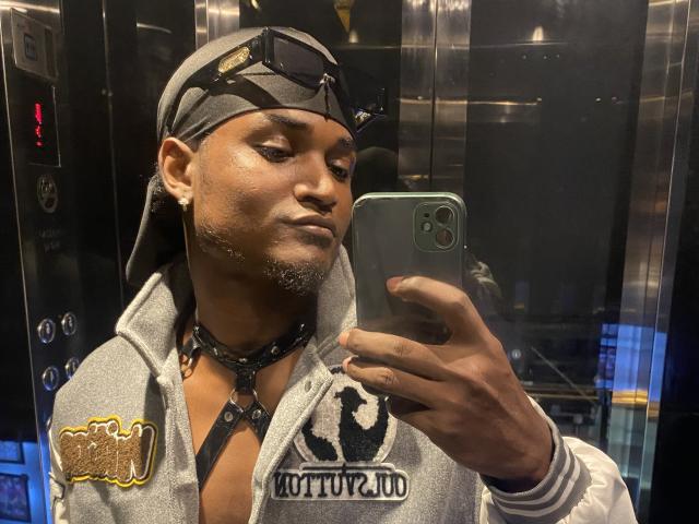 JamesMakony prend un selfie dans un ascenseur, portant un sweat à capuche gris avec le mot "Buttuh" dessus, un collier noir et un bandeau gris avec des lunettes de soleil. Son visage est sérieux..