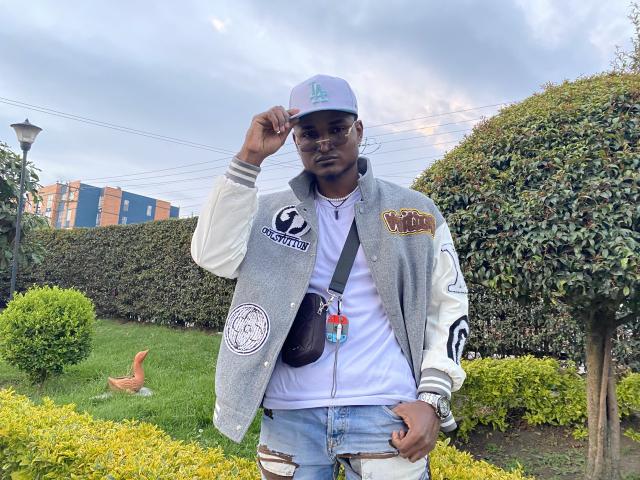 JamesMakony, dark-skinned, wearing a gray varsity jacket with patches, white t-shirt, blue jeans, and a white cap, standing in a green yard with a brown satchel.