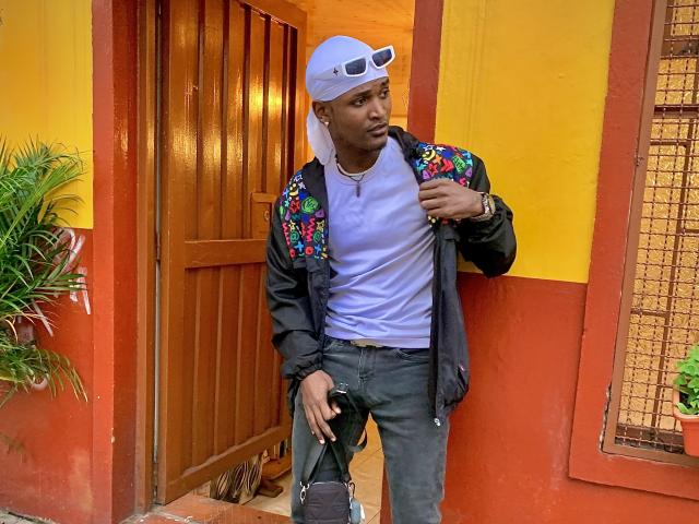 JamesMakony, wearing a white cap, black jacket with colorful embroidery, white tee, and jeans, holding a bag, standing by a wooden door, looking to the side, looking cool.