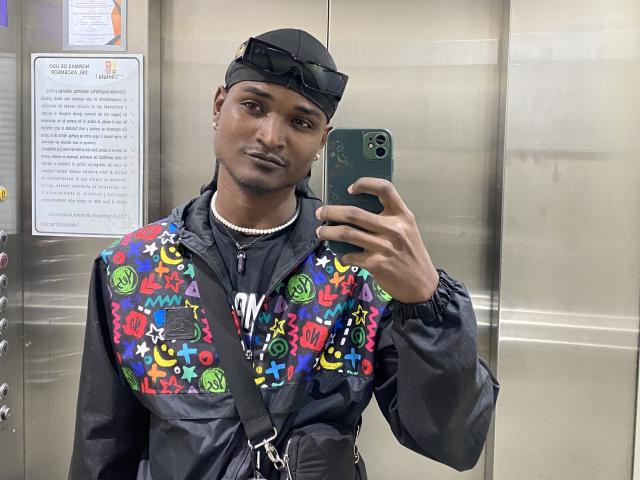JamesMakony with dark skin, black cap, black jacket, colorful graffiti vest, black shirt, necklace, holding green phone, taking selfie, slight smile, mirror reflection, elevator, strap across chest.