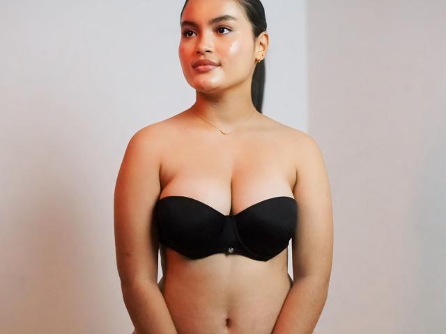MiiaPalmer with tan skin, dark hair in a ponytail, wearing a black strapless bra, showcasing her boobs, a small necklace, and a subtle smile in a simple room.