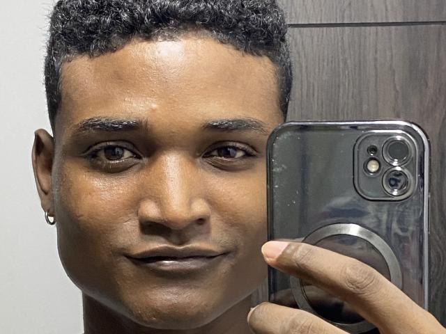 JamesMakony, avec des cheveux noirs et bouclés courts, une peau brune, un petit anneau d'oreille, tenant un iPhone argenté, souriant légèrement, des yeux marrons, regardant la caméra, fond avec une texture en bois..