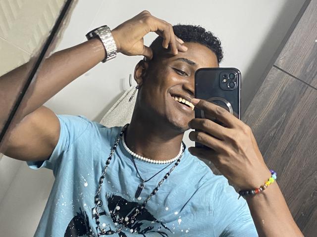 JamesMakony with dark skin, smiling, wearing a blue t-shirt, pearl necklace, beaded bracelet, and silver watch, taking a selfie with a black iPhone in a bathroom.