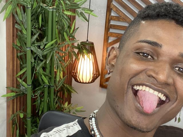 James Makony, avec sa peau foncée, ses cheveux courts et bouclés, et son collier noir, sort sa langue rose et sourit. Une plante de bambou et une lampe suspendue se trouvent en arrière-plan..