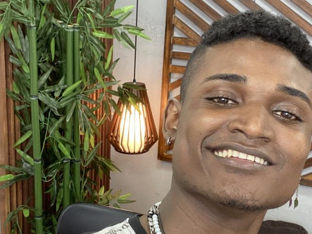 James Makony, ein dunkelhäutiger Mann mit kurzen, lockigen schwarzen Haaren, lächelt mit weißen Zähnen, hat einen kleinen Bart, trägt eine Perlenkette und hat eine Bambuspflanze neben sich stehen..