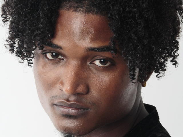 James Makony hat dunkles, lockiges Haar, braune Augen und einen ernsten Gesichtsausdruck. Sein Gesicht hat einen leichten Teint, mit sichtbaren Poren und einem kleinen Bart. Sein schwarzes Hemd ist sichtbar..