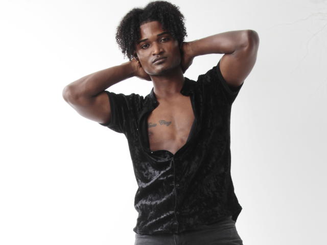JamesMakony with dark skin, curly black hair, hands behind head, black velvet shirt, chest tattoo, exposed nipples, black pants, white background, serious expression, standing, full-body shot.