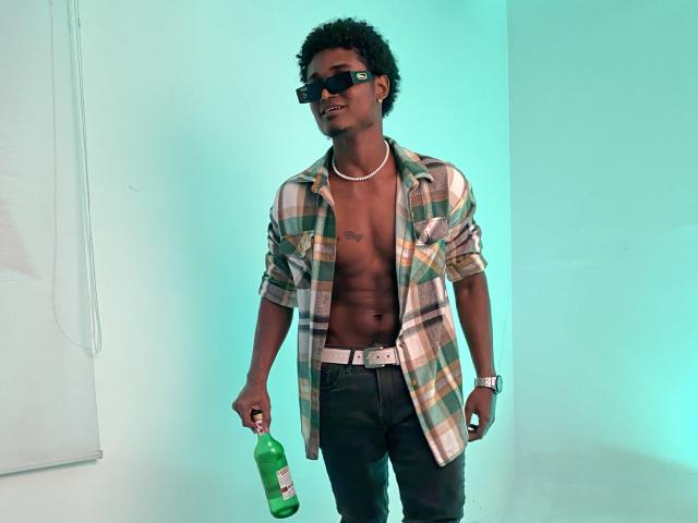JamesMakony, shirtless, wearing black pants, plaid shirt, black sunglasses, necklace, holding green bottle, standing, muscular, dark skin, curly hair, white belt, blue-green light.