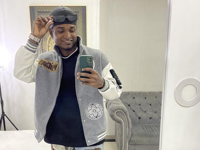 JamesMakony with a dark skin tone, wearing a gray varsity jacket, black shirt, and gray hat, holding a green phone, smiling, and adjusting his hat, standing in front of a mirror.