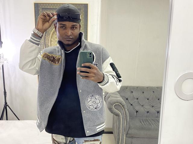JamesMakony, dark-skinned, wearing a gray varsity jacket, black shirt, and backward cap, holding a green phone, standing in front of a mirror, looking at the camera.