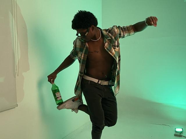 JamesMakony, shirtless, green plaid shirt, dark pants, white belt, white sneakers, holding green bottle, dancing, black curly hair, green light, right leg raised, left arm extended.
