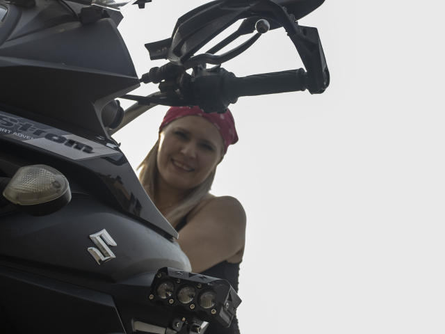 TinaCharter with blonde hair, red bandana, smiling up at a black motorcycle with a "D" logo and headlight, her bare shoulders visible. Motorcycle in foreground.