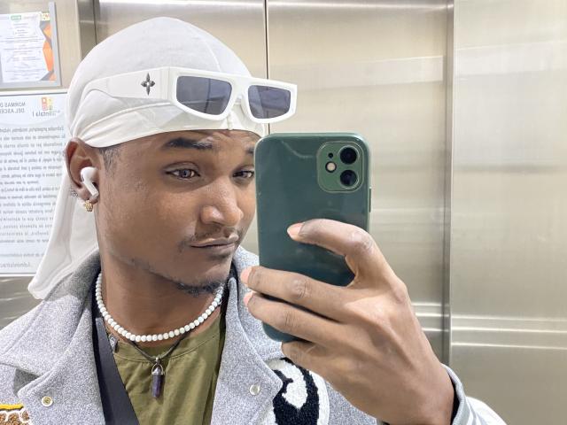 JamesMakony, à la peau foncée, portant un foulard blanc sur la tête, des lunettes de soleil blanches, une chemise verte, une veste grise, un collier de perles, tenant un iPhone vert, prenant un selfie, avec des écouteurs blancs..
