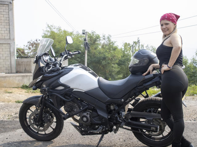 TinaCharter with ass, tits, and a smile, wearing a red bandana, black tank top, and black pants, stands next to a black and silver motorcycle with a helmet.