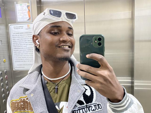 James Makony mit einem weißen Kopftuch, einer Sonnenbrille darauf, einer Perlenkette, einem grünen Telefon, einer grauen Jacke und weißen Ohrringen, der ein Selfie in einem Aufzug macht, lächelnd mit dunkler Haut und Bart..