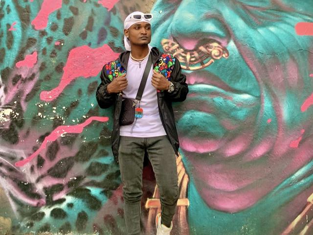 JamesMakony stands against a graffiti wall, wearing a white cap, black jacket with colorful embroidered shoulders, white tee, green pants, and a crossbody bag, looking serious.