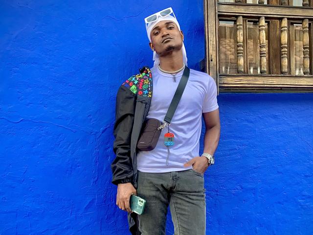 JamesMakony, standing against a bright blue wall, wearing a white cloth on head, white tee, dark jacket with colorful embroidery, gray jeans, holding a green phone, looking up.