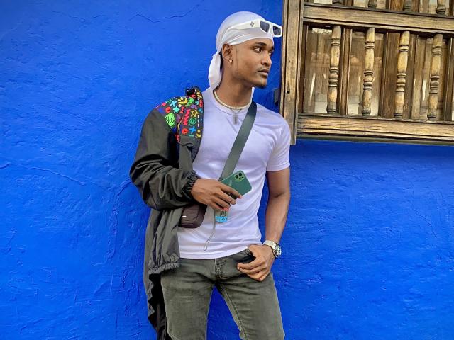 JamesMakony, with a medium brown ass, wears a white cap, grey jacket, colorful shoulder bag, white tee, green jeans, holding a phone, against a bright blue wall.