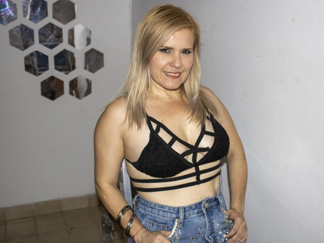 TinaCharter with blonde hair, smiling, wearing a black strappy bralette showing her tits and blue high-waisted jeans, standing in a room with hexagonal wall mirrors.