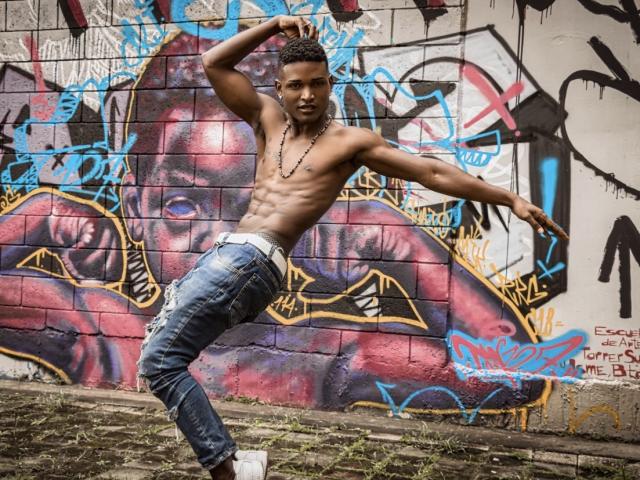 JamesMakony ohne Hemd, mit trainierten Bauchmuskeln, trägt blaue Jeans und eine schwarze Halskette und tanzt mit erhobenen Armen und schwingenden Hüften. Er steht auf einem Kopfsteinpflasterweg vor einer Graffiti-Mauer..