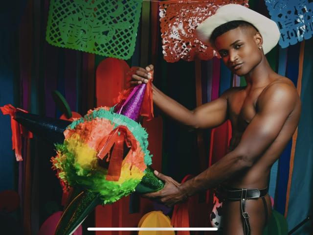 JamesMakony, torse nu, portant un chapeau de cowboy blanc, tenant du papier de cire coloré et un pompon vert, et portant une ceinture noire avec un renflement, a une carrure musclée, de peau foncée et attrayante..