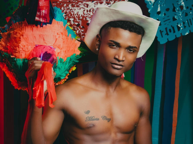 JamesMakony, senza camicia, indossa un cappello da cowboy bianco, tiene in mano dei colorati ritagli di carta (papel picado), ha un tatuaggio sul petto che dice "Love Pig", è muscoloso, ha la pelle marrone, i capelli sono corti e ricci, e guarda la fotocamera..