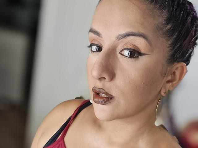 AndreaSharick has medium brown skin, dark brown eyes with winged eyeliner, glossy brown lipstick, and braided purple hair. She's wearing gold hoop earrings and a red top.