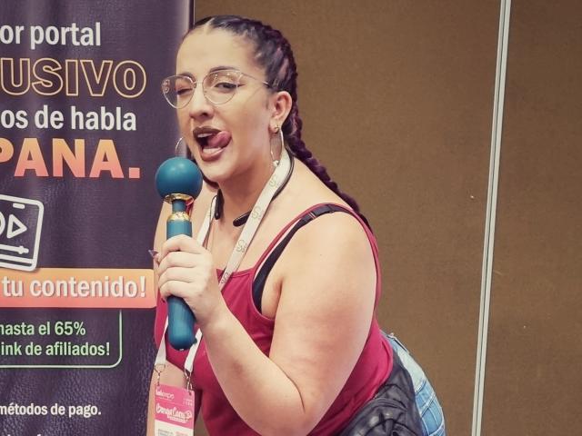 Andrea Sharick, met gevlochten haar, een bril en haar tong uit, houdt een blauwe microfoon vast, draagt een roze lanyard en een rood tanktopje, en laat op de foto haar achterwerk zien..