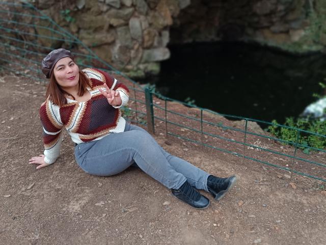 Louise, con cabello castaño, busto grande, vistiendo un suéter de punto, pantalones grises y botas negras, sentada en la tierra, haciendo el gesto de la paz y sonriendo, llevando un gorro gris..