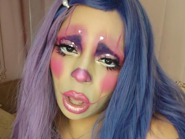 Chili, with blue hair, is making a facial expression with glossy pink lips, heavy pink and purple eyeshadow, and semen on her face.