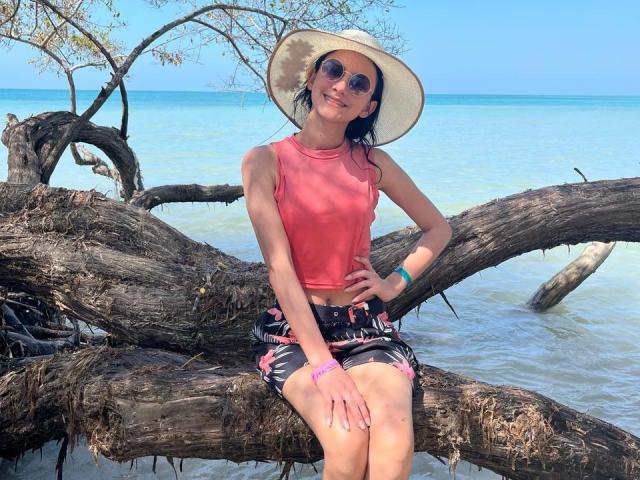 KateDavies with small tits, wearing a pink sleeveless top, black floral skirt, sunhat, and sunglasses, sitting on a fallen tree at a beach, with clear blue water.